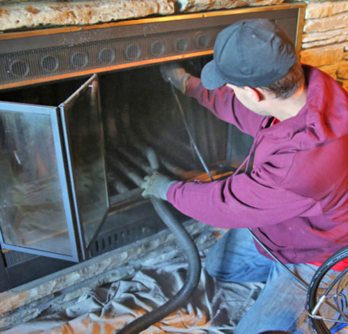 A person wearing gloves and a cap uses a vacuum hose to clean the inside of a fireplace.