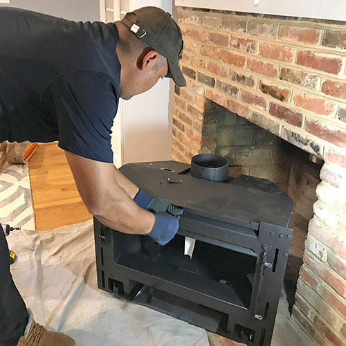 A person wearing gloves and a cap is installing or servicing a black wood stove in front of a brick fireplace.