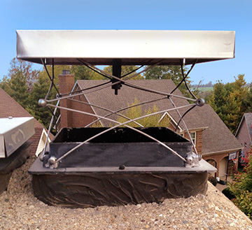 A metal chimney cap with a spring-loaded mechanism is installed on top of a brick chimney, with residential rooftops visible in the background.