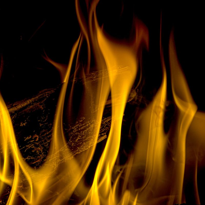 Close-up of bright yellow and orange flames with a charred wooden log partially visible in the background against a dark backdrop.