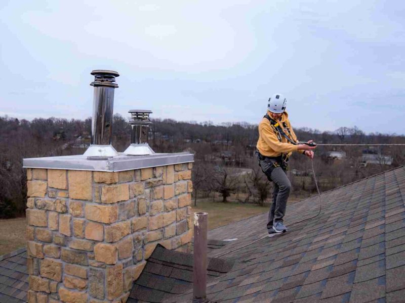 A person wearing safety gear and a helmet stands on a sloped roof next to a stone chimney, holding a rope, with trees and houses visible in the background.