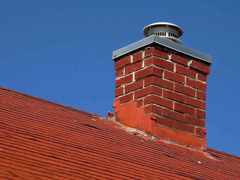 A red brick chimney with a metal cap sits on a red shingled roof against a clear blue sky.
