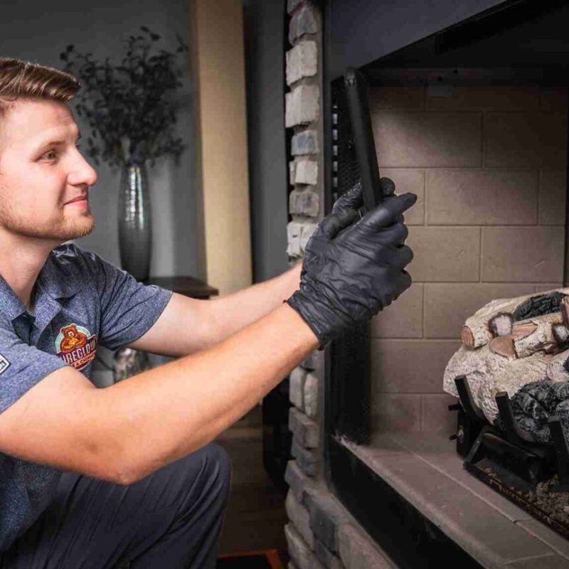 A technician wearing black gloves inspects the inside of a fireplace, holding a flashlight and kneeling near the hearth.