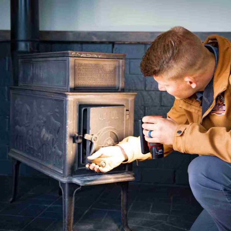 A person in a brown jacket kneels to open the door of a black cast-iron wood stove, holding a can in one hand as sunlight shines on the stove’s interior.