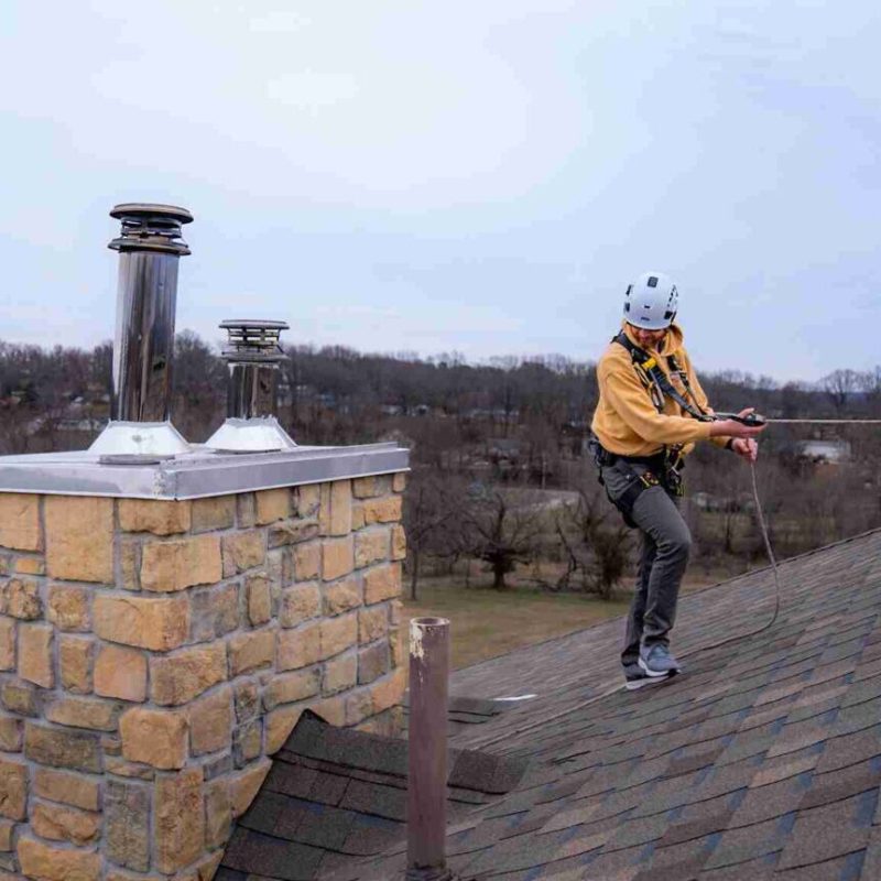 A person wearing safety gear and a helmet secures a rope while standing on a sloped roof next to a stone chimney on a cloudy day, with trees and houses visible in the background.
